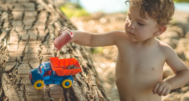 Niño jugando al aire libre. Niño, echamos arena en el camión rojo. Juegos de calle para niños. Un niño jugando con una máquina en el tronco grande.