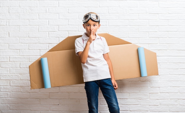 Niño jugando con alas de aeroplano de cartón en la espalda mostrando un signo de boca cerrada