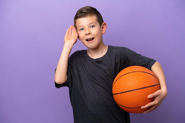 Niño jugando baloncesto aislado en un fondo morado escuchando algo poniendo la mano en la oreja