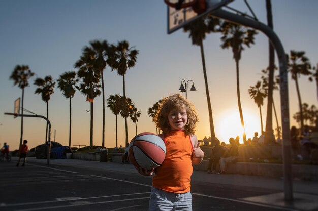 Niño jugando baloncesto con pelota de baloncesto al aire libre en la puesta de sol. Pulgares hacia arriba.