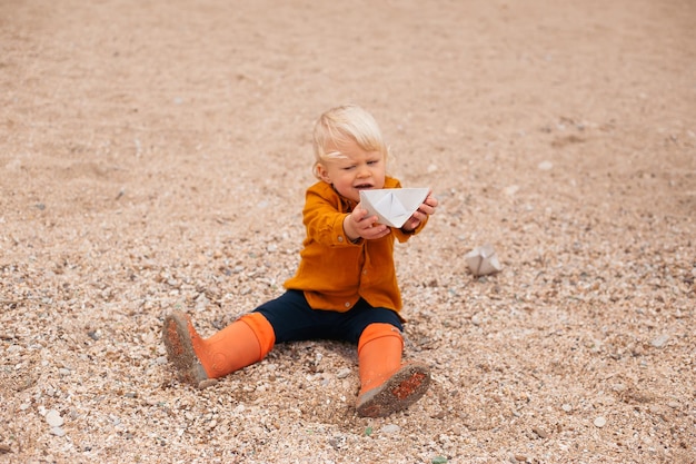 Niño jugando con un barco de papel en la playa de arena cerca del mar en otoño o verano