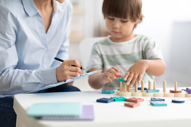 Niño jugando juguete de madera lógico en la oficina de psicólogo mujer especialista tomando notas selectivas