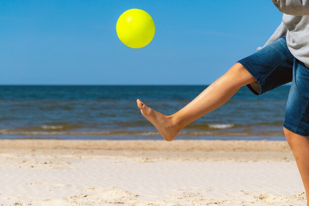 Niño jugando con una pelota de playa en la arena junto al agua del mar en un día soleado