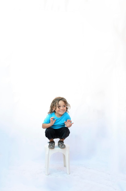 Foto niño jugando en silla vestido con fondo azul y blanco cabello largo y rizado sonriendo