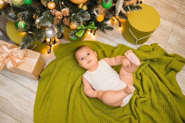Un niño juguetón con un traje blanco yace sobre una manta de punto verde debajo de un árbol de Navidad con regalos con una copia del espacio