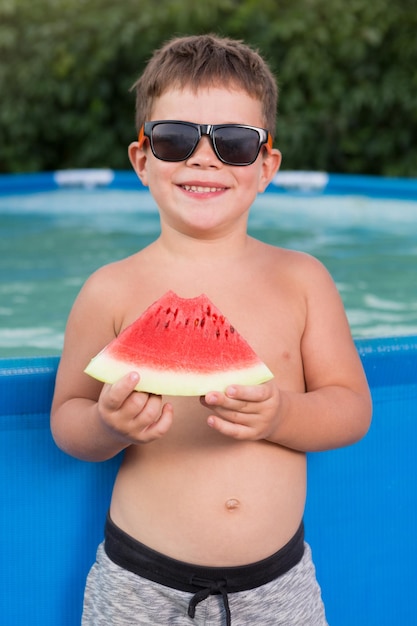 Niño junto a la piscina azul sosteniendo un trozo de sandía roja y riendo de verano