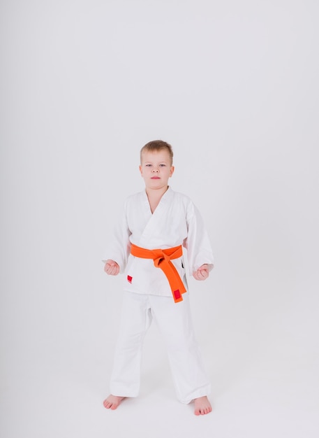 Niño con un kimono blanco con un cinturón naranja se encuentra en una pose en una pared blanca con una copia del espacio