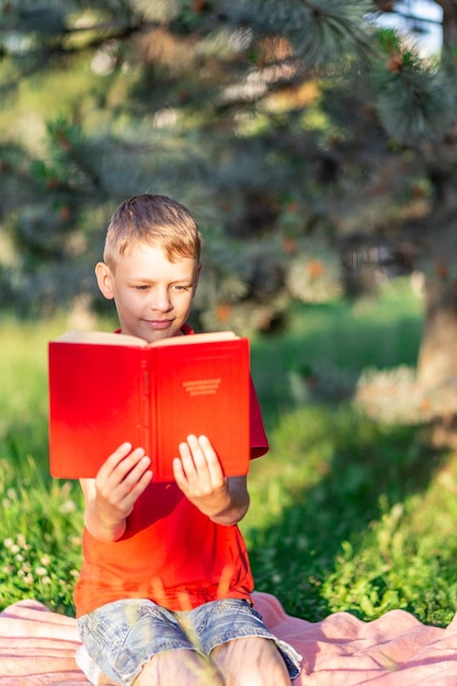 un niño lee un libro rojo sentado en una manta contra un fondo de vegetación