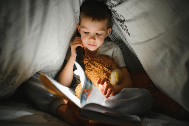 Niño leyendo un libro en la cama. Los niños leen por la noche. Niño pequeño con libros de cuentos de hadas en el dormitorio. Educación para niños pequeños. Cuento antes de dormir por la noche. Niño lindo debajo de una manta en una habitación oscura con lámpara.