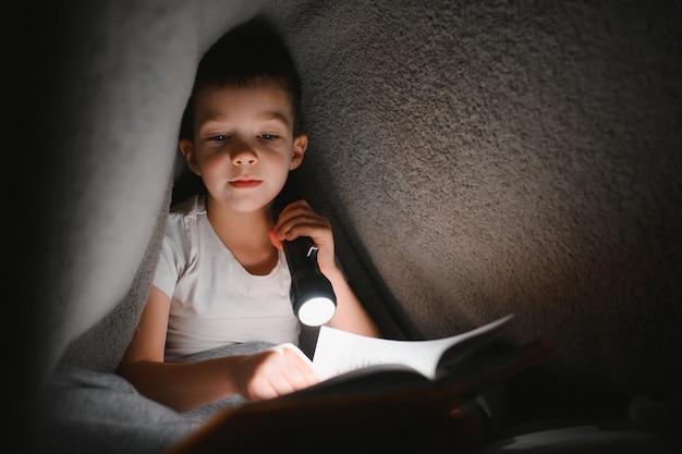 Niño leyendo un libro en la cama. Los niños leen por la noche. Niño pequeño con libros de cuentos de hadas en el dormitorio. Educación para niños pequeños. Cuento antes de dormir por la noche. Niño lindo debajo de una manta en una habitación oscura con lámpara.