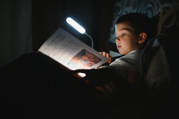 Niño leyendo un libro en la cama Los niños leen por la noche Niño pequeño con libros de cuentos de hadas en el dormitorio Educación para niños pequeños Cuento para dormir por la noche Niño lindo debajo de una manta en una habitación oscura con lámpara