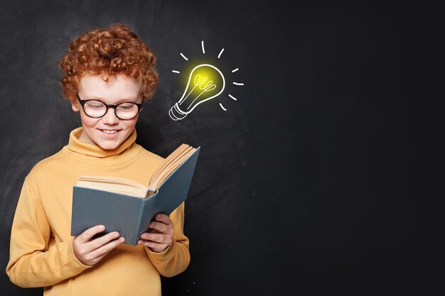 Foto niño leyendo un libro divirtiéndose y con la idea de una bombilla en el fondo de la pizarra en el aula