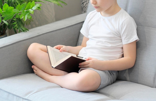 Niño leyendo un libro en el sofá con las piernas cruzadas foto clara en primer plano gris y blanco sin rostro visible