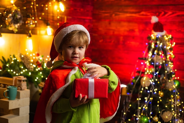 Niño lindo abriendo un regalo de Navidad. Niño alegre vestido como Santa Claus. Un niño con gorro de Papá Noel ayuda con el regalo de Navidad en una caja roja.