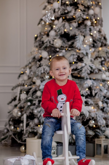 Niño lindo con accesorios de Navidad sentado en caballo de madera delante del árbol de Navidad