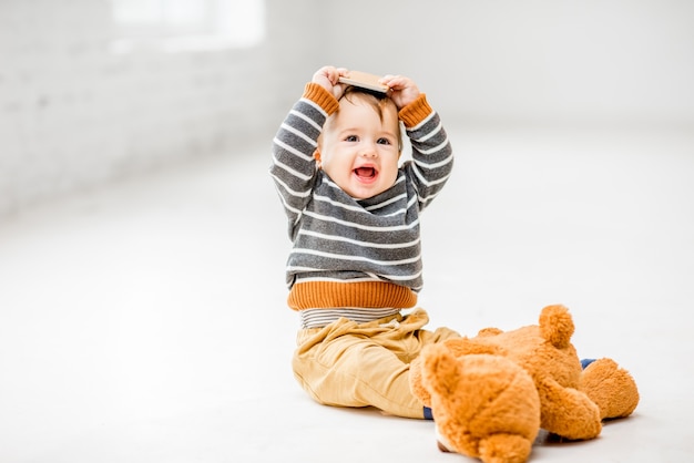 Niño lindo y alegre jugando con el teléfono inteligente sentado en el interior en el piso blanco