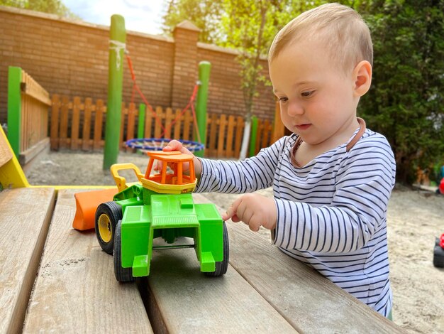 Un niño lindo de un año y medio juega con un coche de juguete en el patio de recreo Adorable niño pequeño jugando con coches y juguetes al aire libre Concepto de estilo de vida de los niños de vacaciones familiares Enfoque selectivo