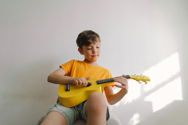 Foto un niño lindo aprende a tocar el ukulele amarillo en un hogar acogedor.