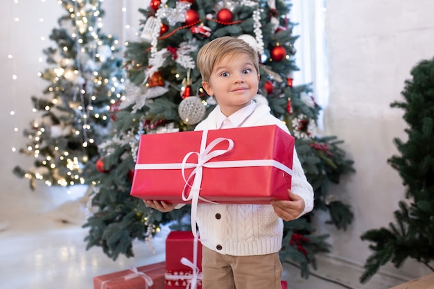 Niño lindo con caja de regalo de Navidad cerca de árboles de Navidad con luces. Feliz navidad y felices fiestas