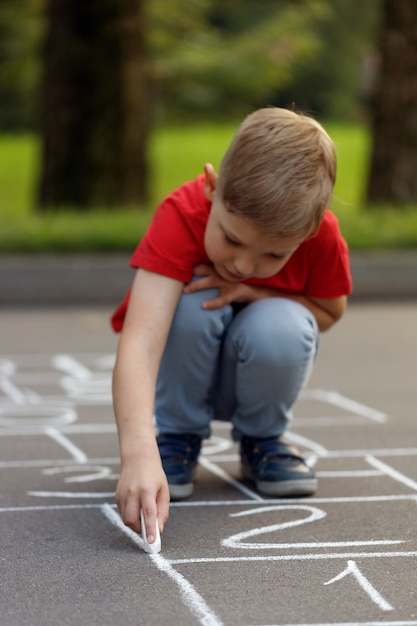 Foto niño lindo en camiseta roja dibujo rayuela con tiza en el patio de recreo.