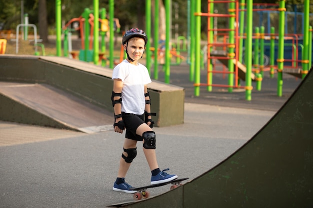 Niño lindo en un casco de pie en un área especial en skatepark y pisar patineta El niño realiza trucos Concepto de actividad deportiva de verano Infancia feliz