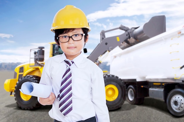 Niño lindo con casco de seguridad y estampado azul frente a un camión de carga de excavadora con tierra en un sitio de construcción