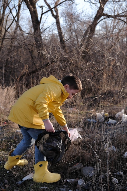 Un niño lindo con chaqueta amarilla y botas de goma está recogiendo basura afuera