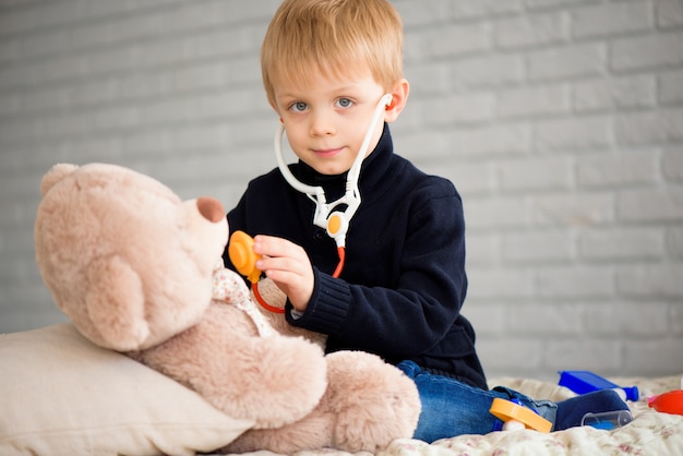 Niño lindo disfrazado de médico jugando con oso de juguete en casa.