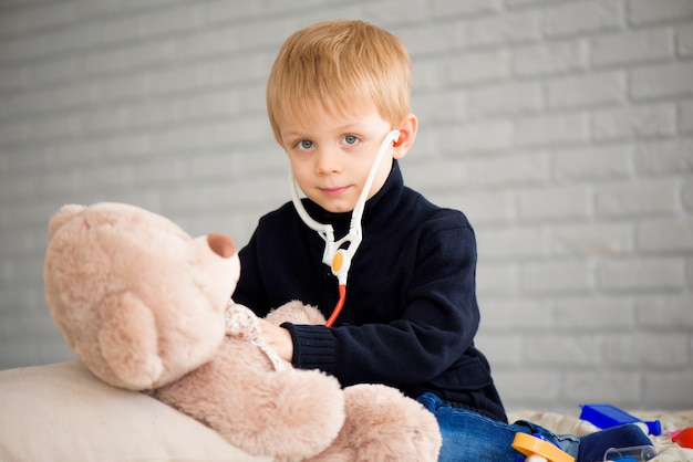 Niño lindo disfrazado de médico jugando con oso de juguete en casa.