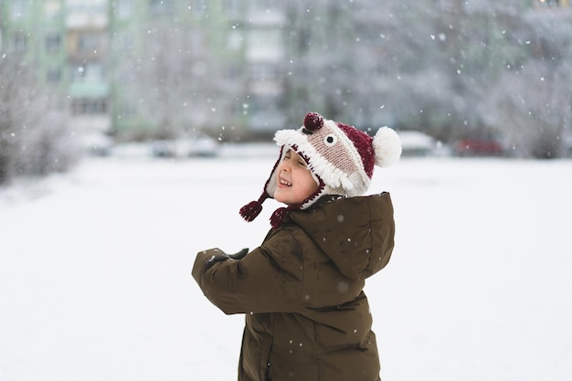 Un niño lindo con un divertido sombrero de invierno camina durante una nevada al aire libre actividades de invierno para niños