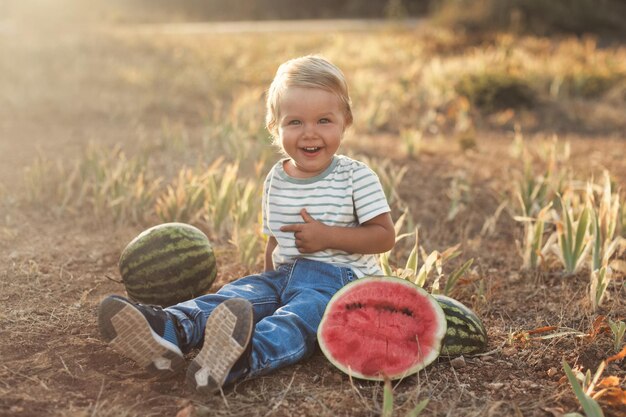 Niño lindo y emotivo riéndose del día soleado sentado en la hierba con sandía fresca