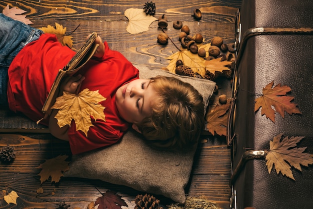 Niño lindo se encuentra en un piso de madera con hojas de otoño y lee el libro. Niño niño se encuentra en un