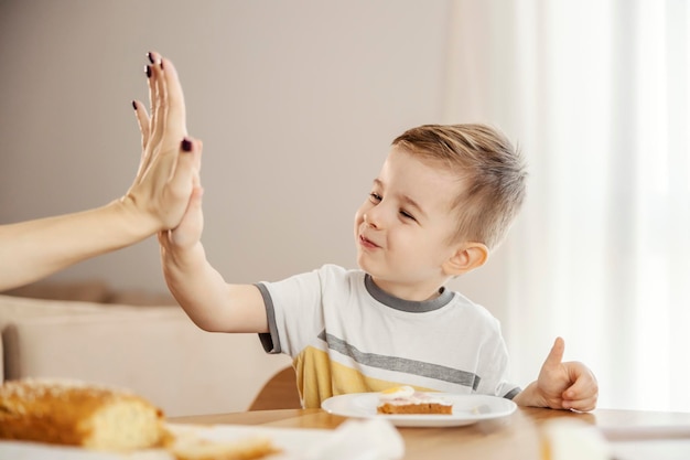 Un niño lindo está desayunando saludablemente y masticando la comida mientras choca los cinco con la madre