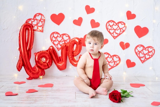 Un niño lindo está sentado con una corbata roja y bragas sobre un fondo de corazones rojos y la inscripción amor concepto de día de san valentín día de san valentín