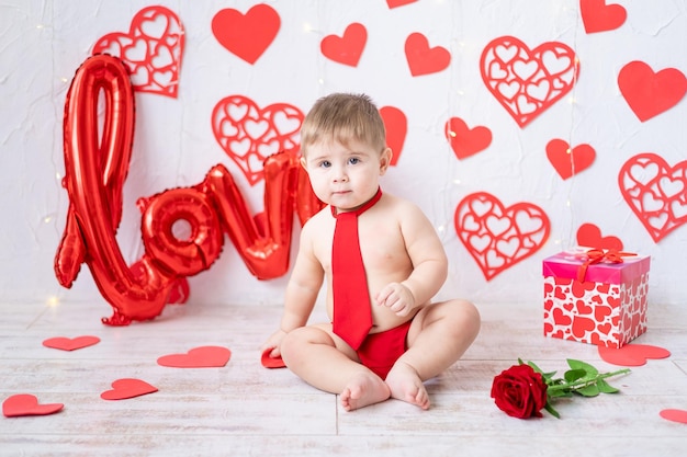 Un niño lindo está sentado con una corbata roja y bragas sobre un fondo de corazones rojos y la inscripción amor concepto de día de san valentín día de san valentín