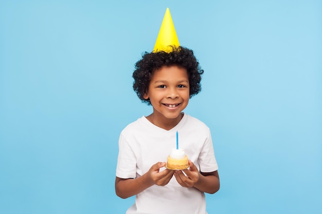 Un niño lindo y feliz con un divertido cono de fiesta en la cabeza sosteniendo un pastel con una vela y sonriendo a la cámara con una cara complacida y satisfecha, expresando pura felicidad infantil. tiro del estudio aislado sobre fondo azul