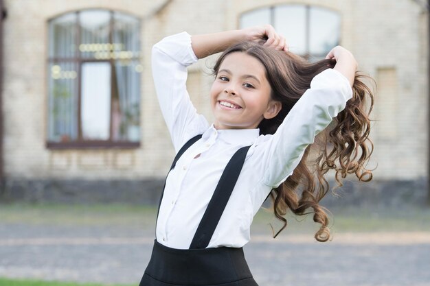 Niño lindo feliz en uniforme escolar arreglar cabello largo ondulado morena en el patio de la escuela al aire libre cuidado del cabello