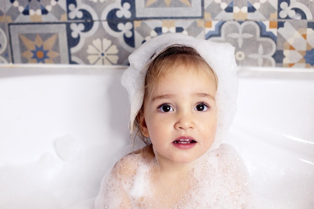 Niño lindo con grandes ojos hermosos sentado en el baño con espuma de baño. Cuidado del bebé, cuidado personal. Copie el espacio. 18-24 meses niña mirando a la cámara