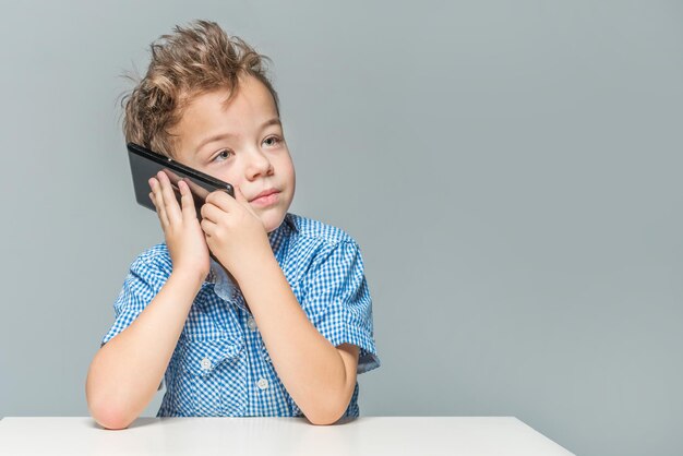 Niño lindo hablando por teléfono mientras está sentado en la mesa sobre un fondo gris aislado
