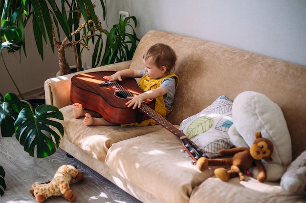 Un niño lindo juega con una gran guitarra acústica en casa en el sofá.