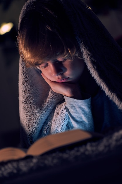 Niño lindo leyendo un libro interesante y apoyándose en la mano mientras se acuesta en una cama cómoda bajo una manta cálida en una habitación oscura en casa