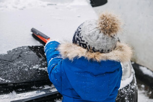 Niño lindo limpiar la nieve del coche en el patio de invierno. Niño pequeño Niño ayudando al padre a limpiar el coche de la nieve y usando la herramienta de cepillo para limpiar el coche.