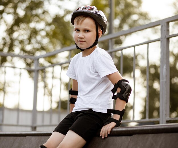 Niño lindo niño niño en un casco sentado en un área especial en skatepark y sosteniendo monopatín Concepto de actividad deportiva de verano Infancia feliz