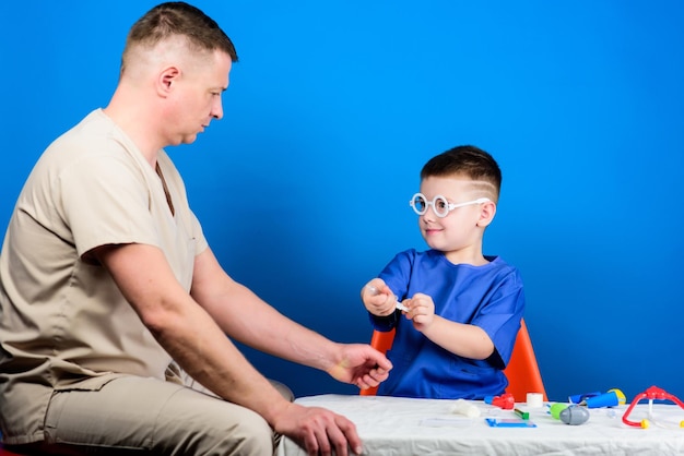 Niño lindo niño y su padre médico Trabajador del hospital Concepto de medicina Niño pequeño doctor sentarse mesa herramientas médicas Tratamiento de enfermedades Papá e hijo dinastía médica Atención médica Examen médico