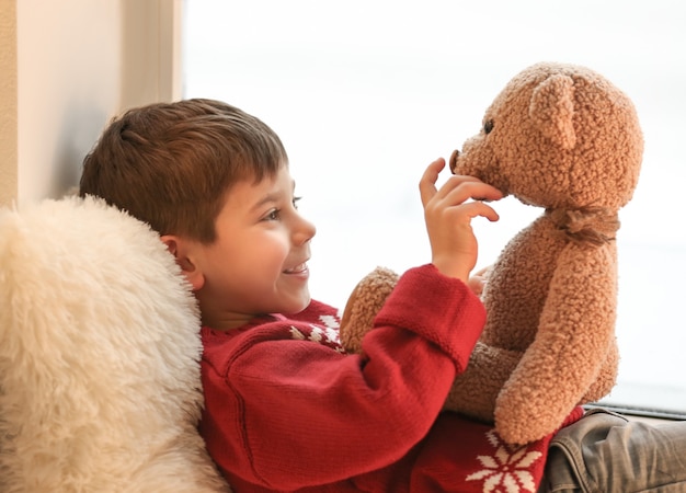 Niño lindo con osito de peluche junto a la ventana en casa