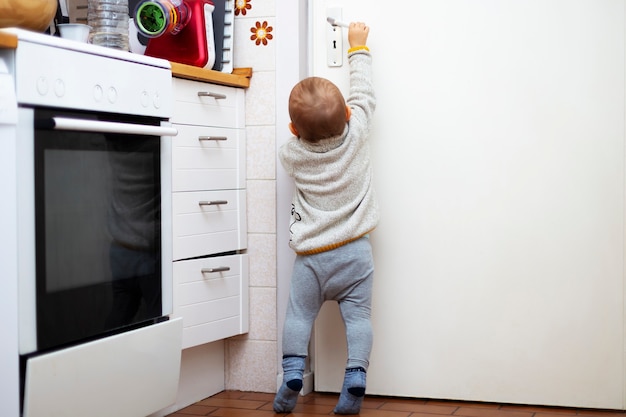 Un niño lindo quiere abrir una puerta en la habitación. Retrato de un bebé tratando de alcanzar una manija de la puerta de la cocina con la mano. Seguridad infantil.