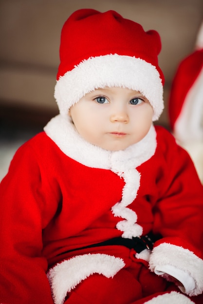 Niño lindo de Santa en suite roja y gorra sentada en un sillón grande cerca del árbol de Navidad