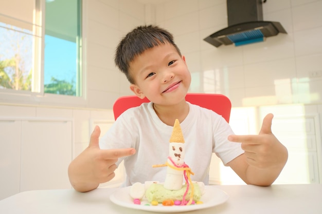 Niño lindo sentado en la cocina haciendo fácil y divertida artesanía comestible Marshmallow Snowman para niños