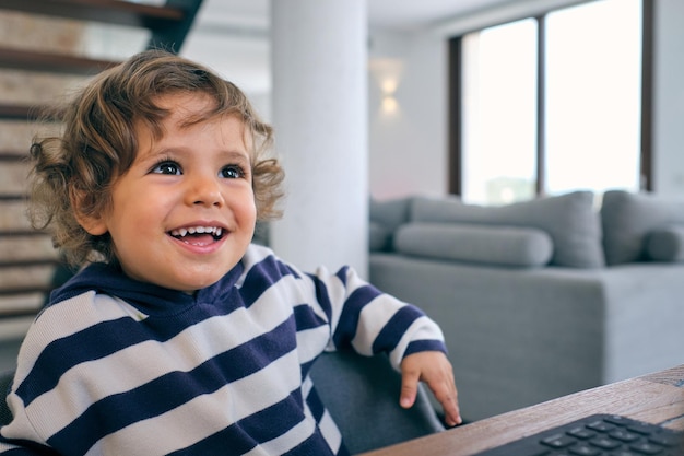 Niño lindo sentado en la mesa con una computadora portátil y mirando hacia otro lado mientras pasa tiempo en la acogedora sala de estar en casa