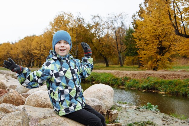 Un niño lindo sentado en una roca junto a un arroyo contra los árboles.